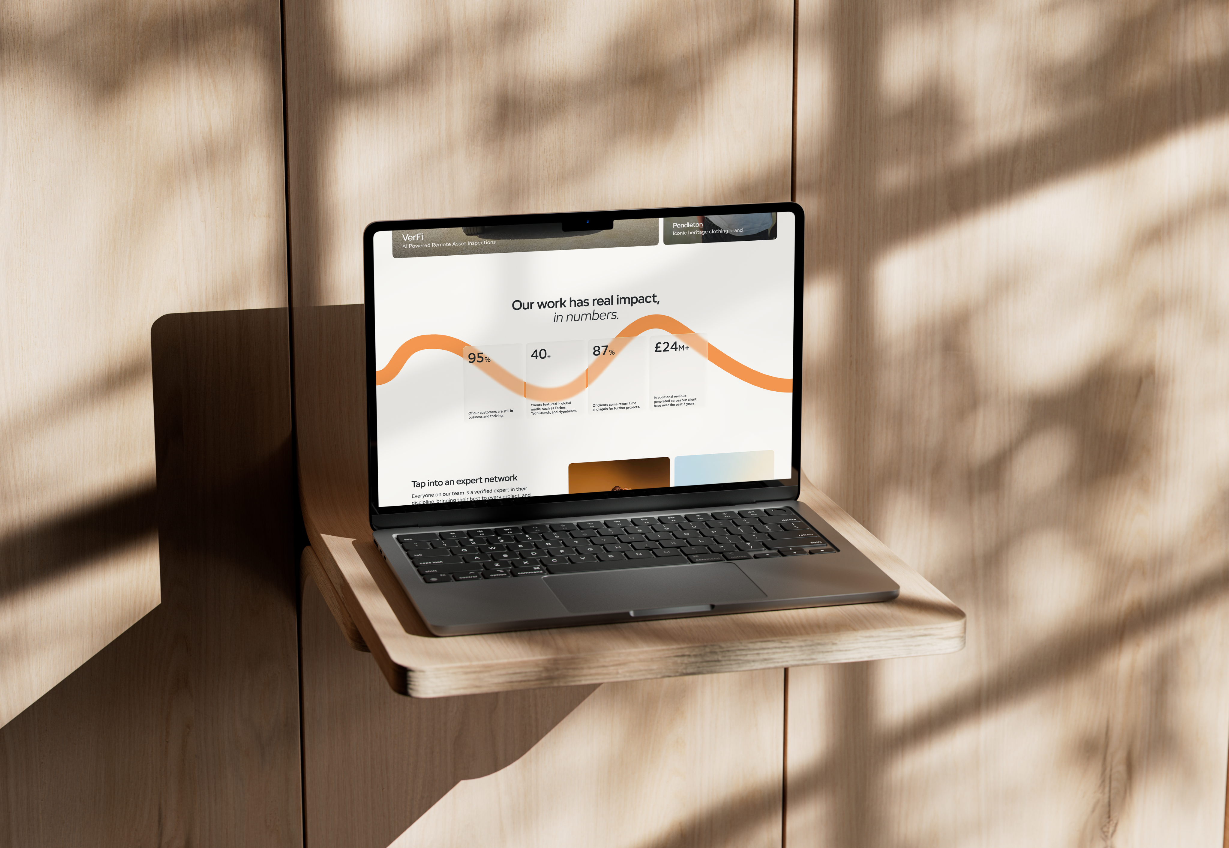 Laptop on a wooden shelf against a sunlit timber wall, displaying a website with an “Our work has real impact” headline and orange impact statistics.