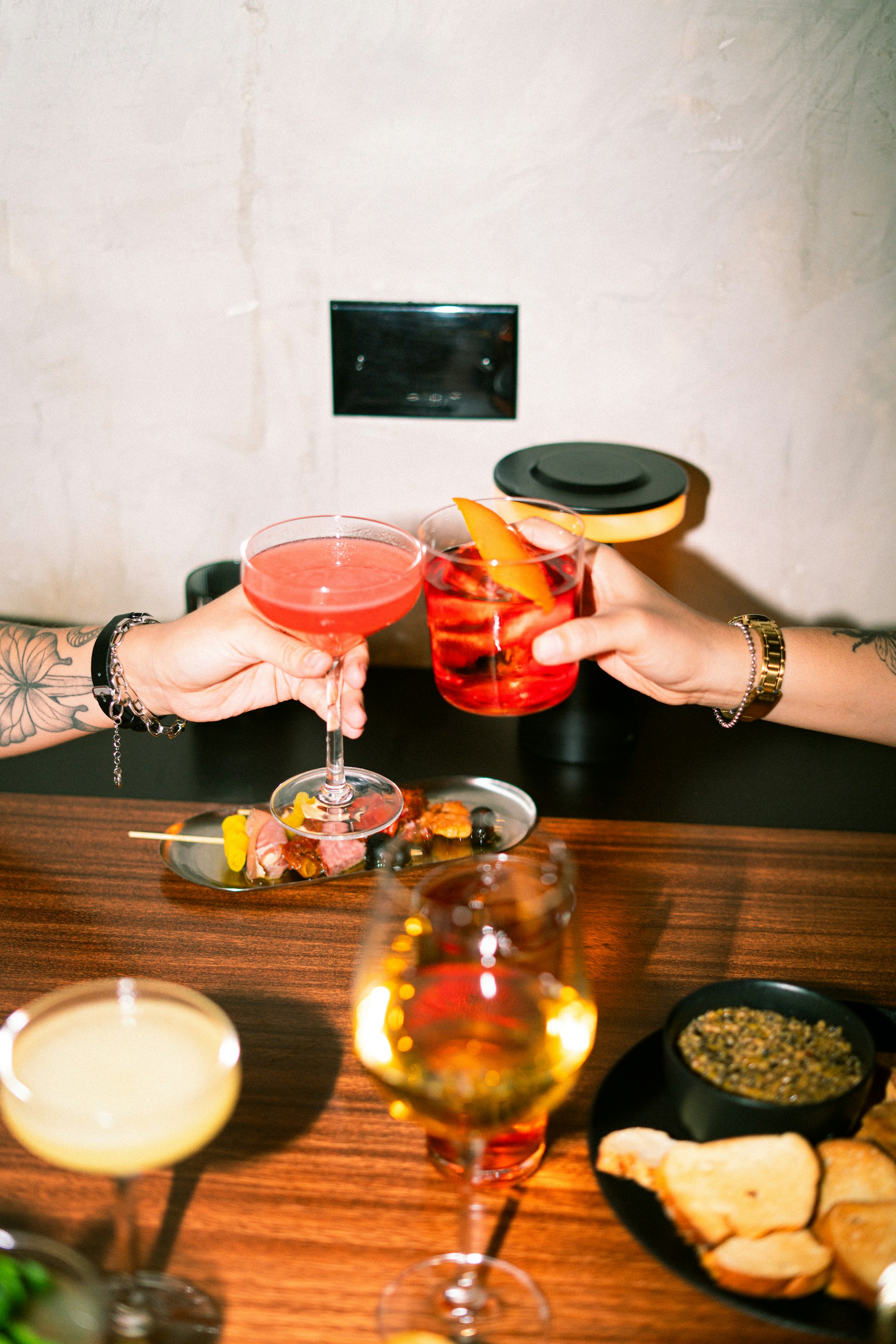 Two hands clinking cocktail glasses over a wooden bar with tapas.